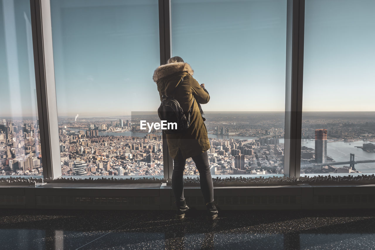 REAR VIEW OF WOMAN LOOKING THROUGH GLASS WINDOW IN CITY