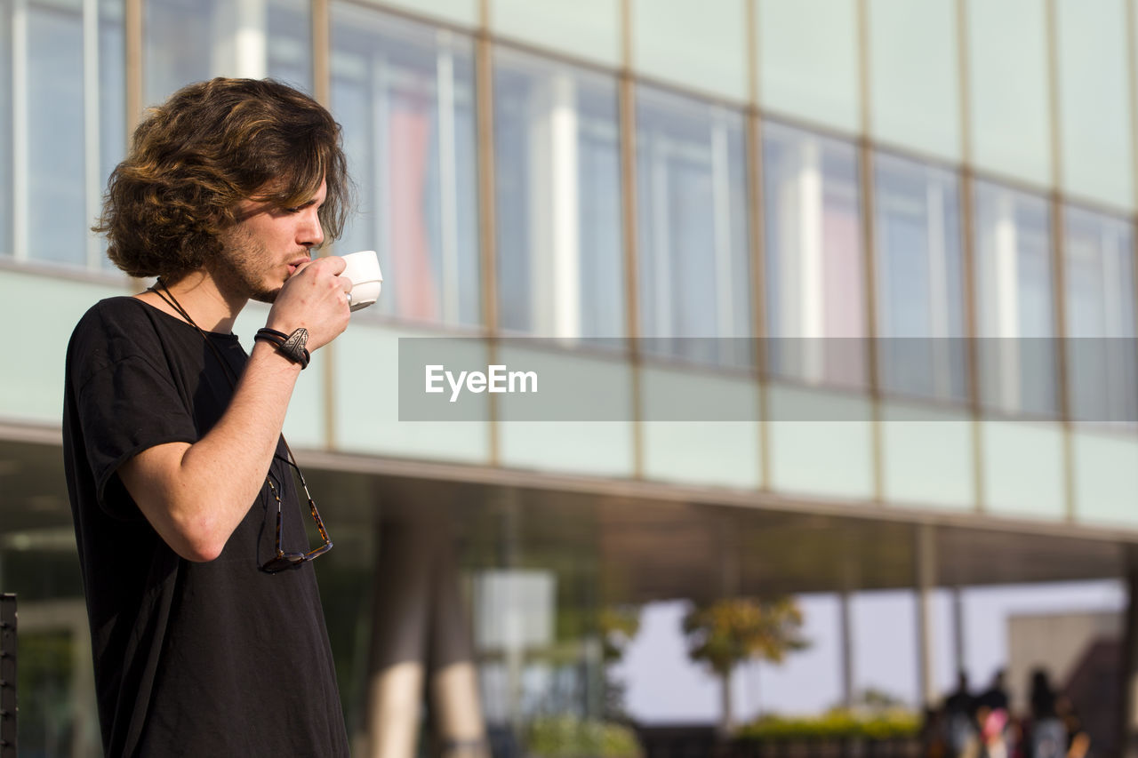 Young man drinking coffee while standing against building