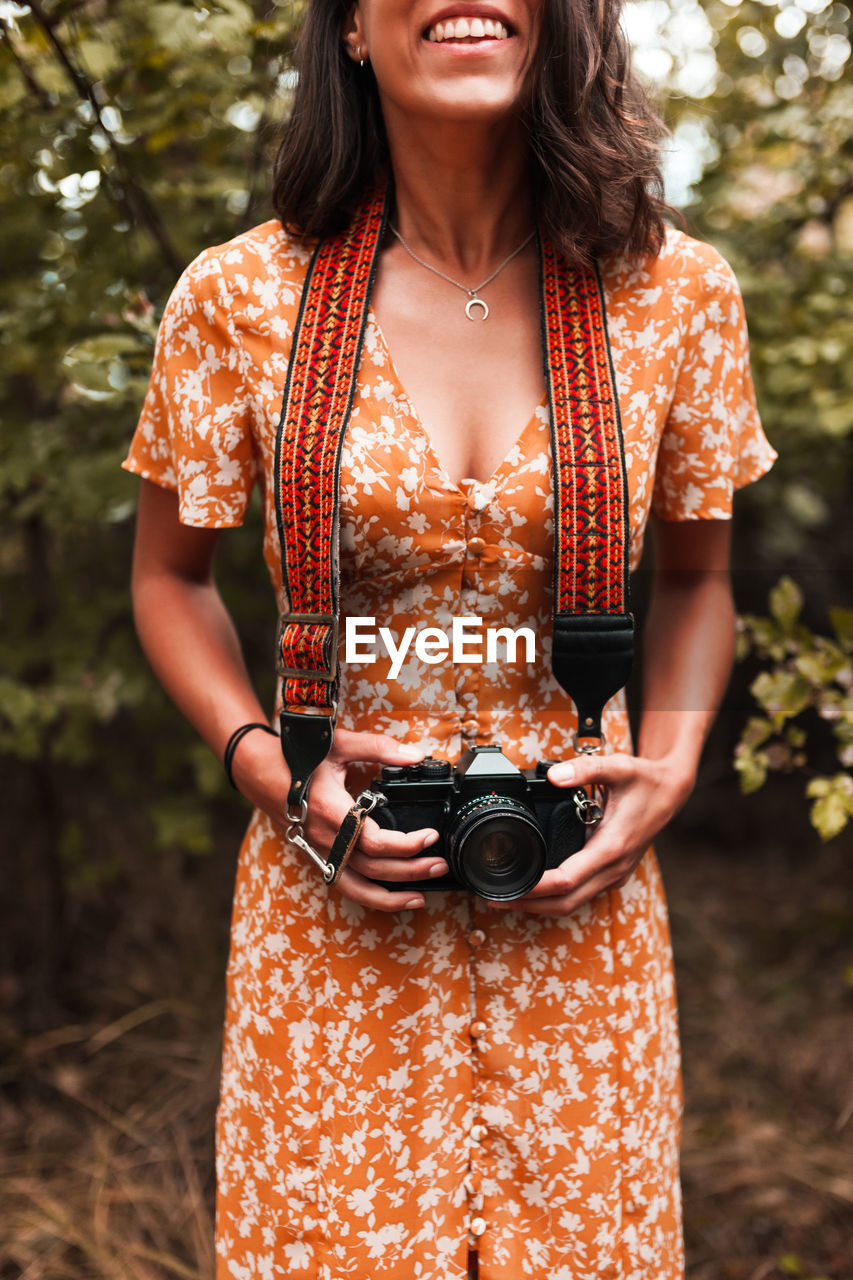 Midsection of young woman with camera standing against tree