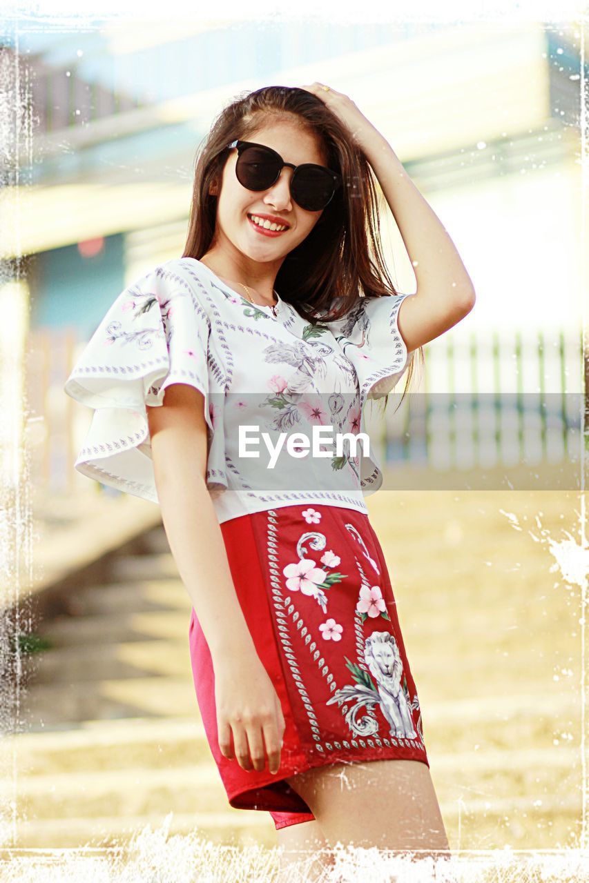 PORTRAIT OF SMILING YOUNG WOMAN WEARING SUNGLASSES STANDING OUTDOORS