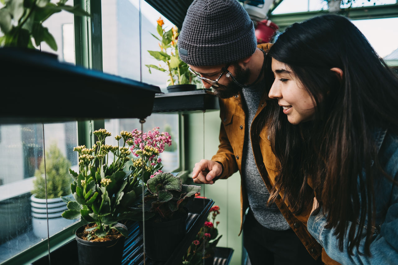 Content stylish couple embracing in greenhouse while standing near shelf and picking blooming potted flower