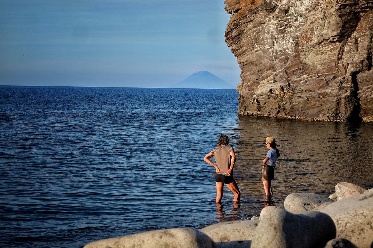 People standing in sea by rock formation against sky