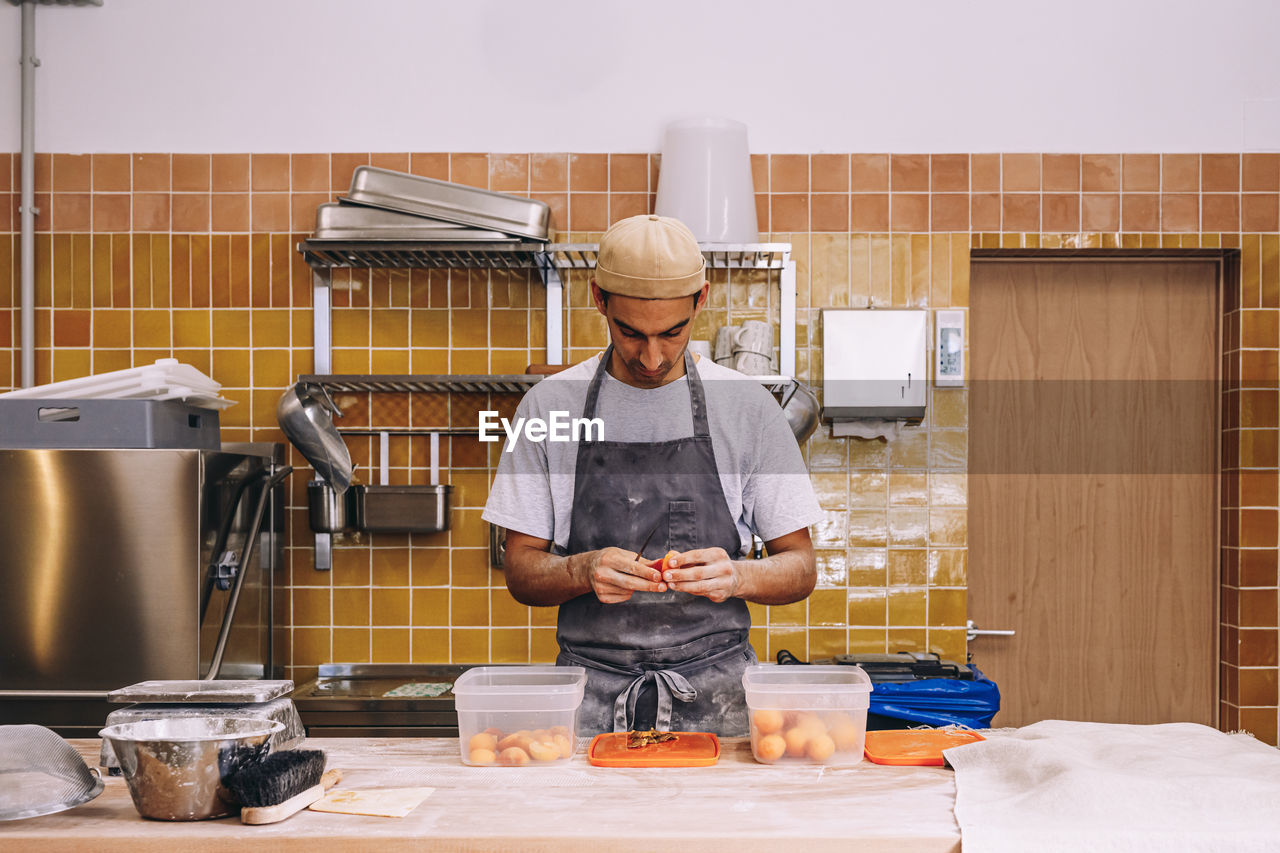 Male baker wearing apron standing at table in bakehouse and preparing fresh fruits for cooking