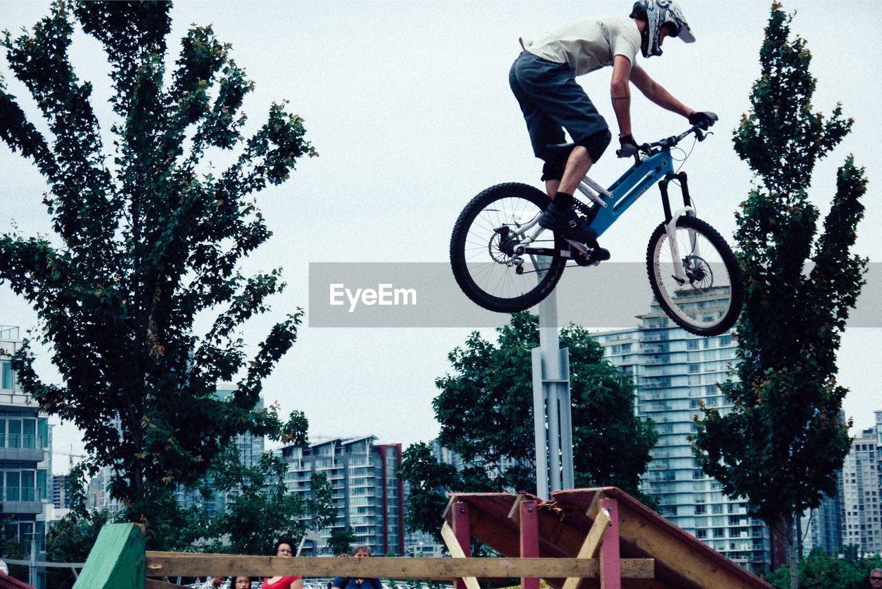 Low angle view of young man jumping on bicycle in city against sky