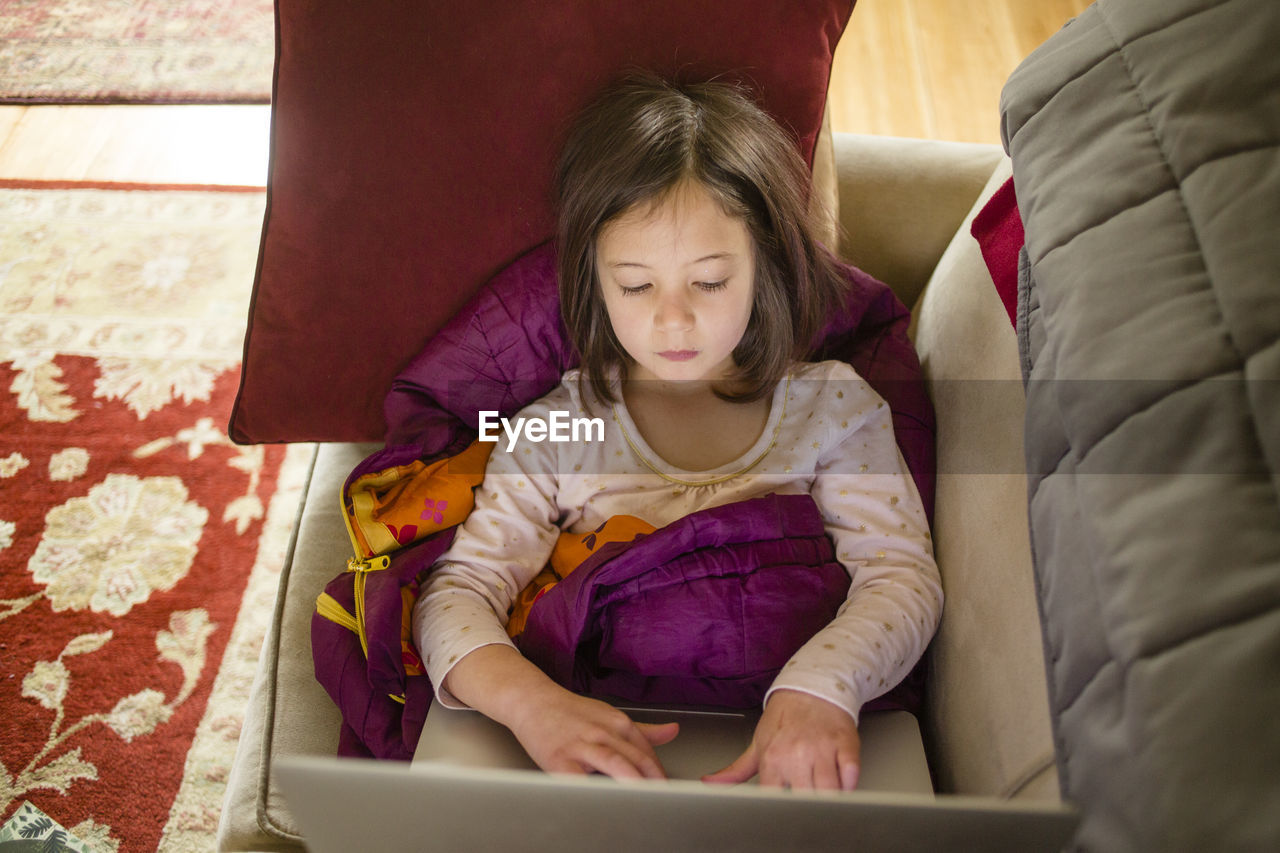 A little girl sits on couch in a pile of blankets working on computer