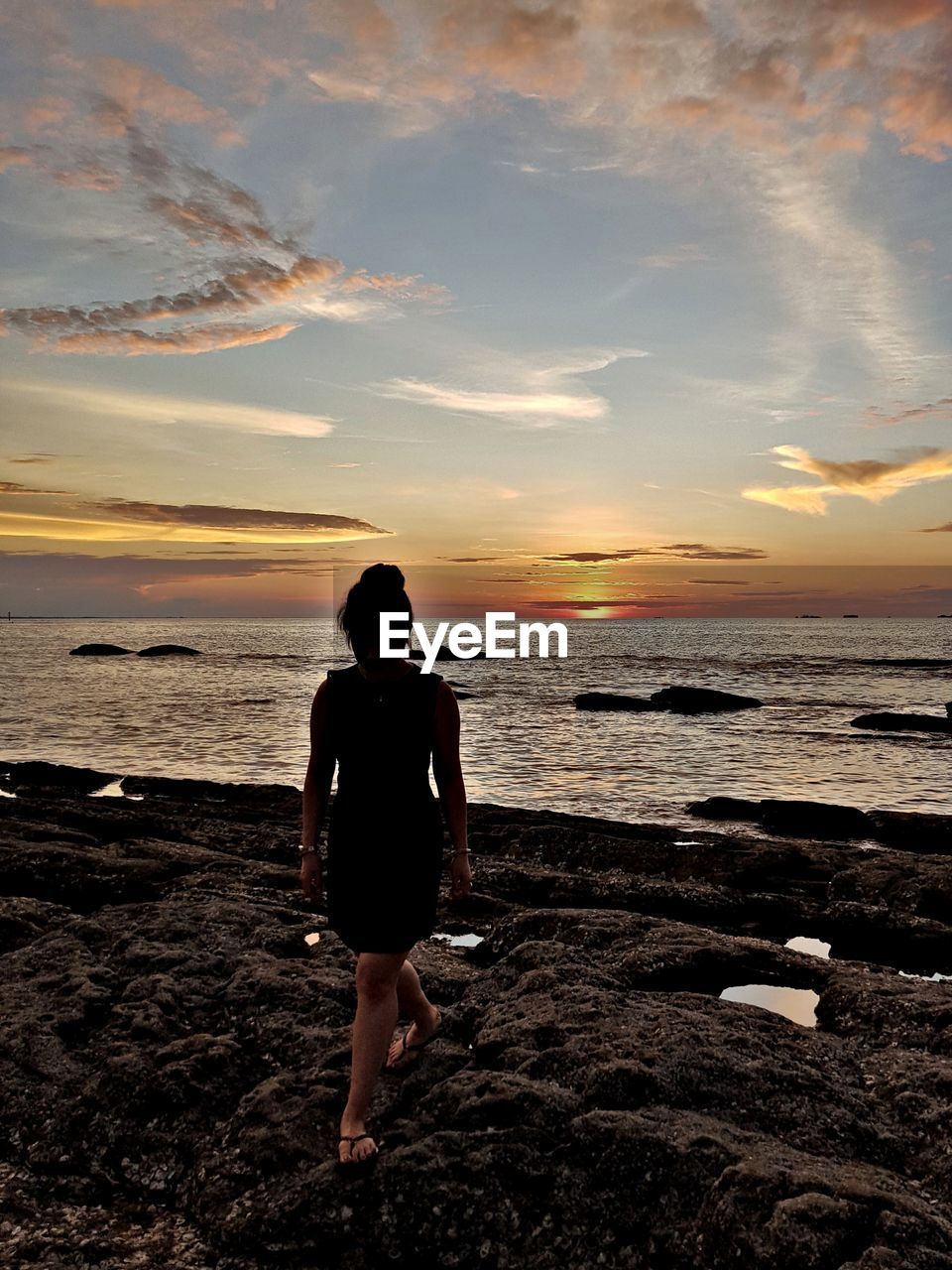 REAR VIEW OF WOMAN STANDING ON BEACH DURING SUNSET