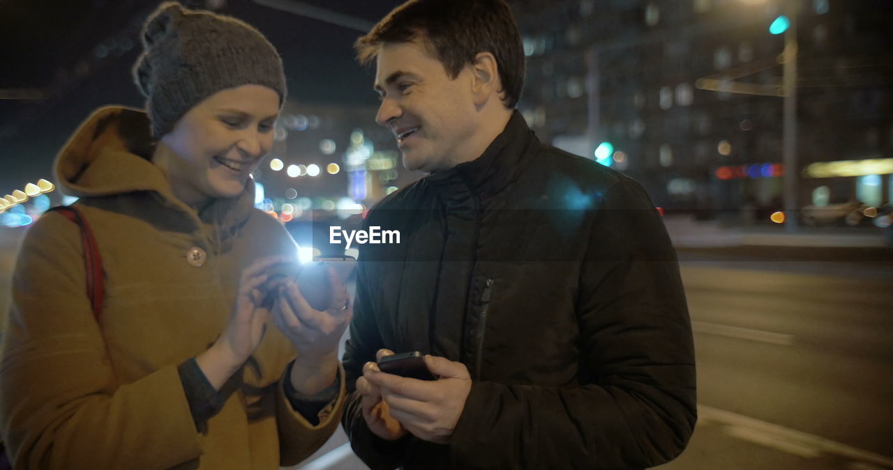 YOUNG MAN HOLDING SMART PHONE WHILE STANDING ON ILLUMINATED CITY