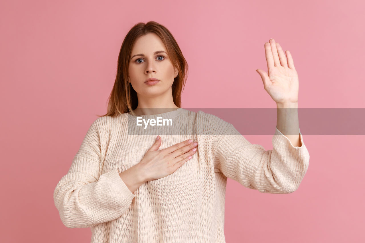 pink, one person, studio shot, portrait, women, adult, colored background, sleeve, finger, waist up, indoors, young adult, looking at camera, standing, hand, pink background, gesturing, brown hair, hairstyle, arm, front view, outerwear, clothing, sign language, limb, female, emotion, photo shoot, long hair, human limb, sweater, casual clothing, facial expression