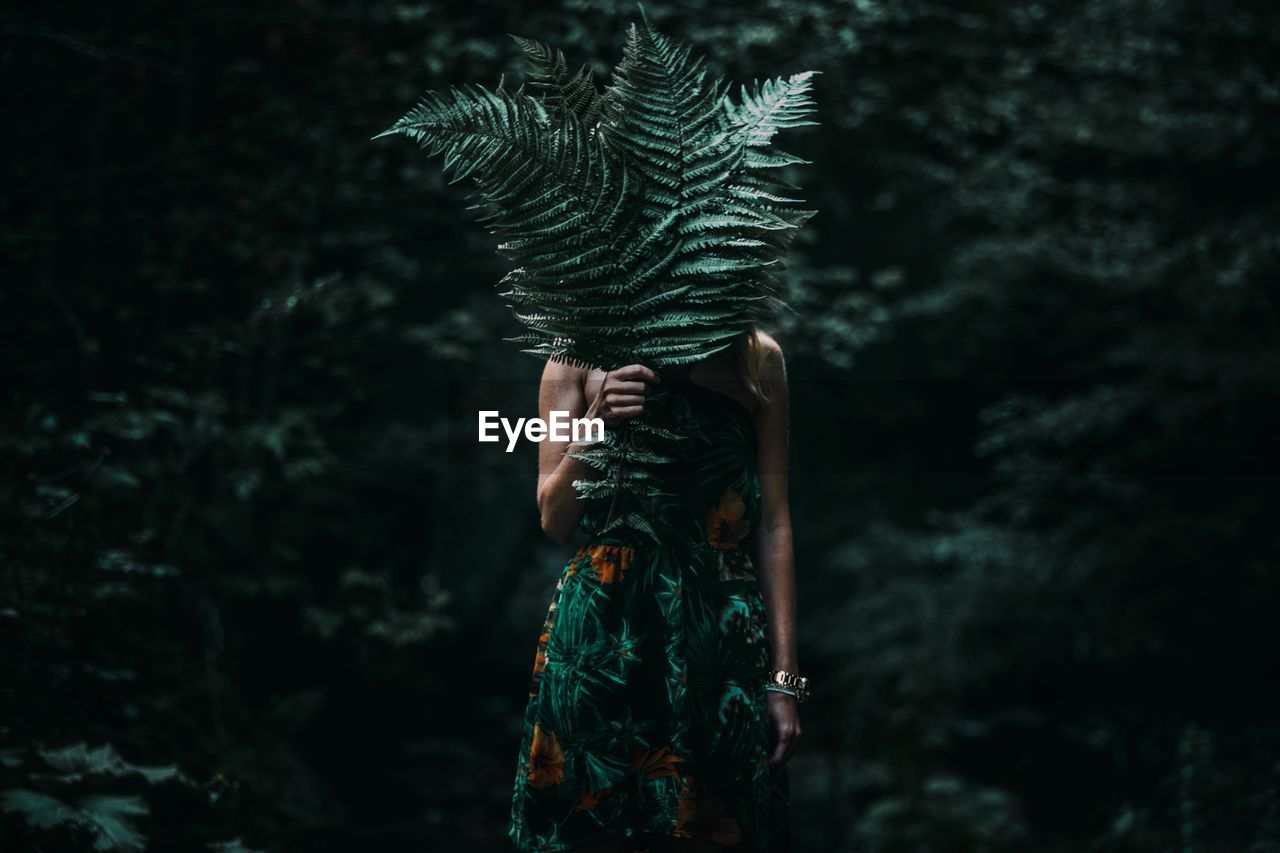 Woman covering face with leaves while standing in forest