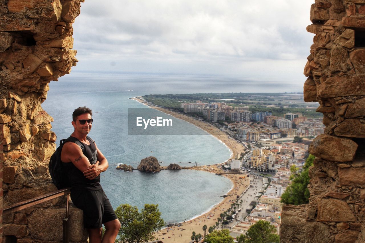 Portrait of man with arms crossed standing by sea against cityscape