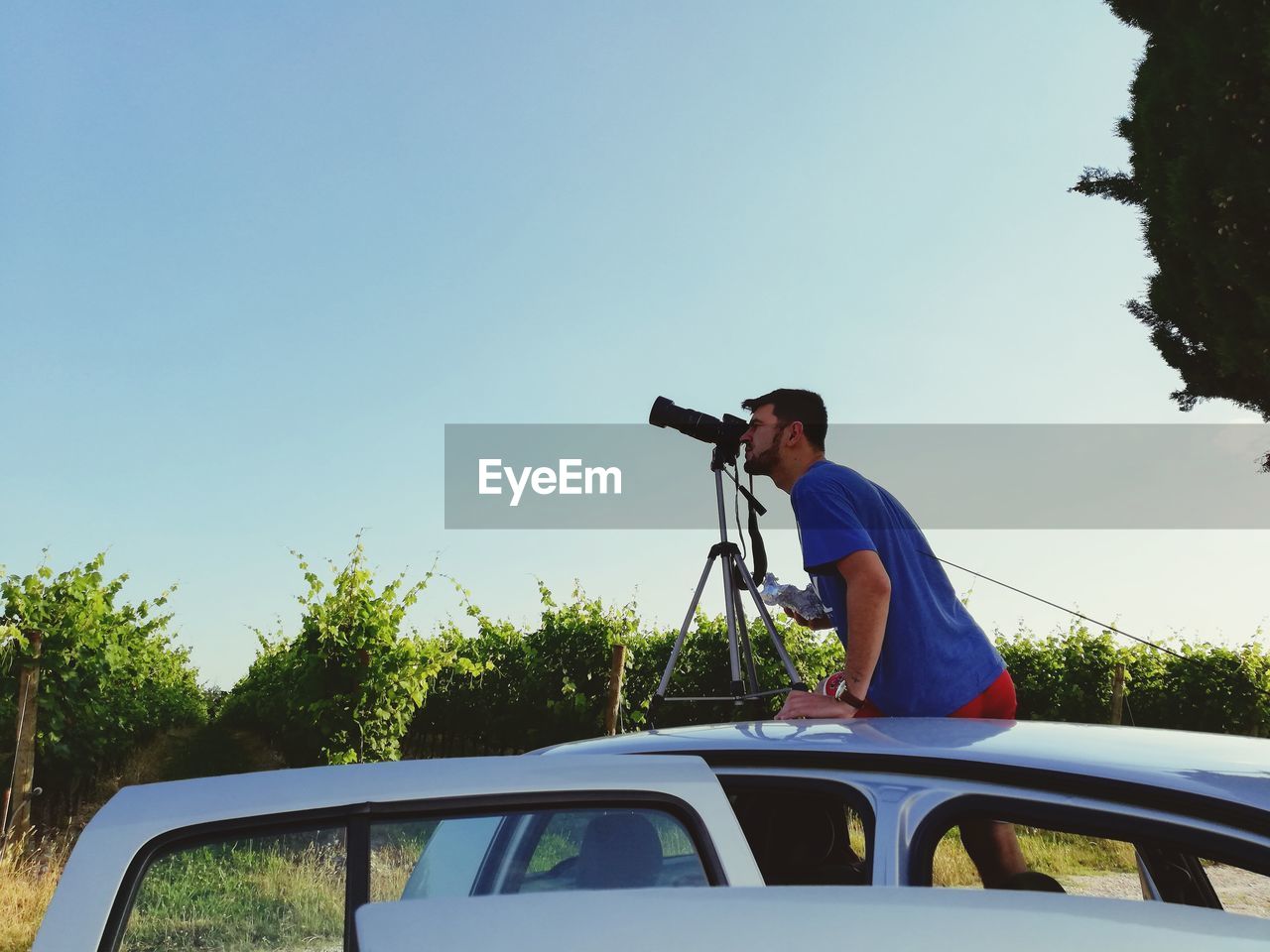 Side view of man photographing on car roof against blue sky