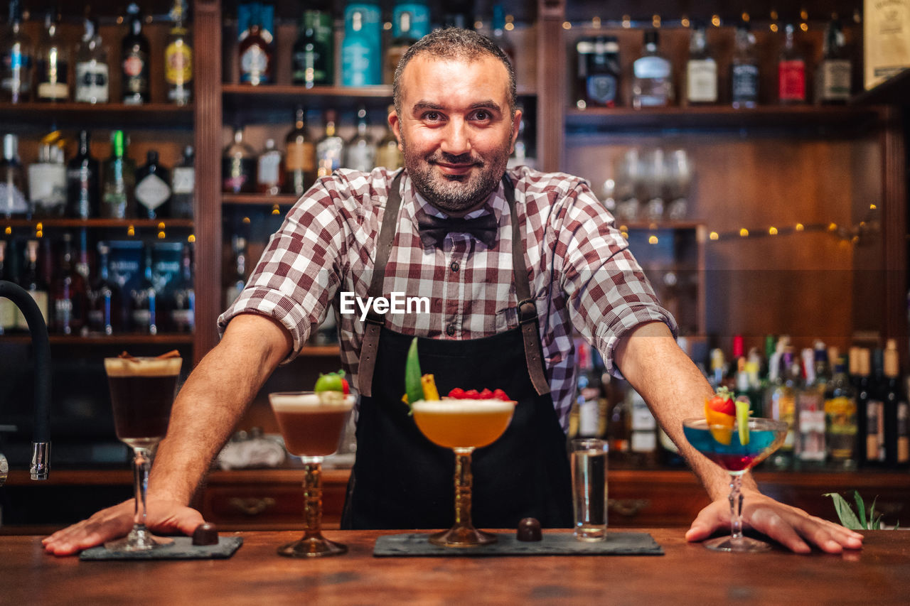 Bearded bartender smiling while standing near glasses of fresh cold sour cocktails at counter in bar looking at camera