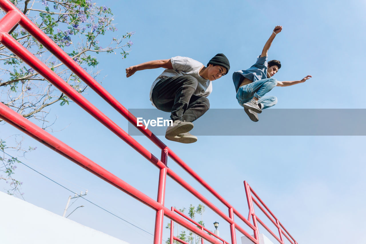 From below fearless male friends jumping above metal railing in city while performing parkour stunt on sunny day