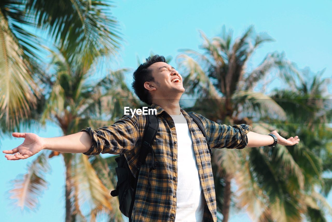 Cheerful man with arms outstretched standing against palm trees