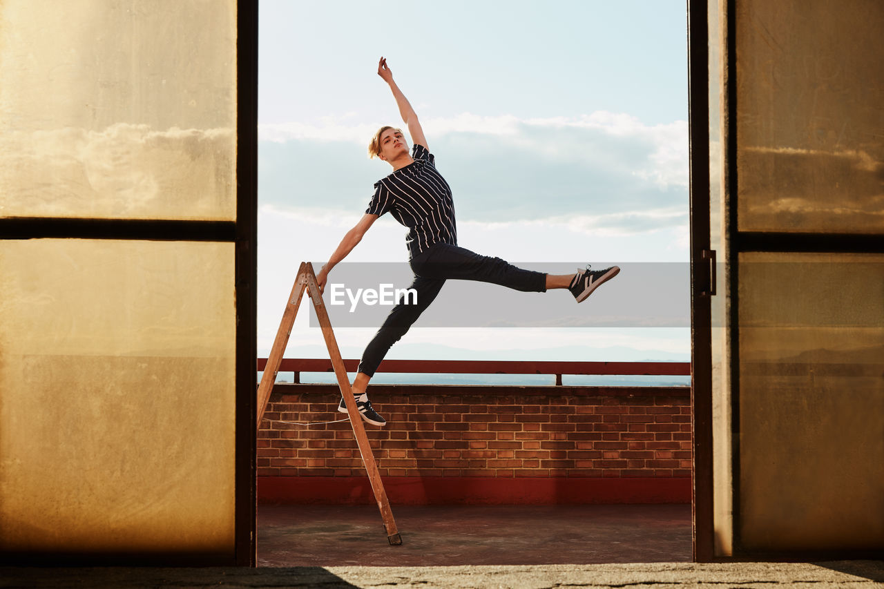 Fashionable elegant young performer standing on one leg on stepladder and raising hand to sky on roof of building on sky background