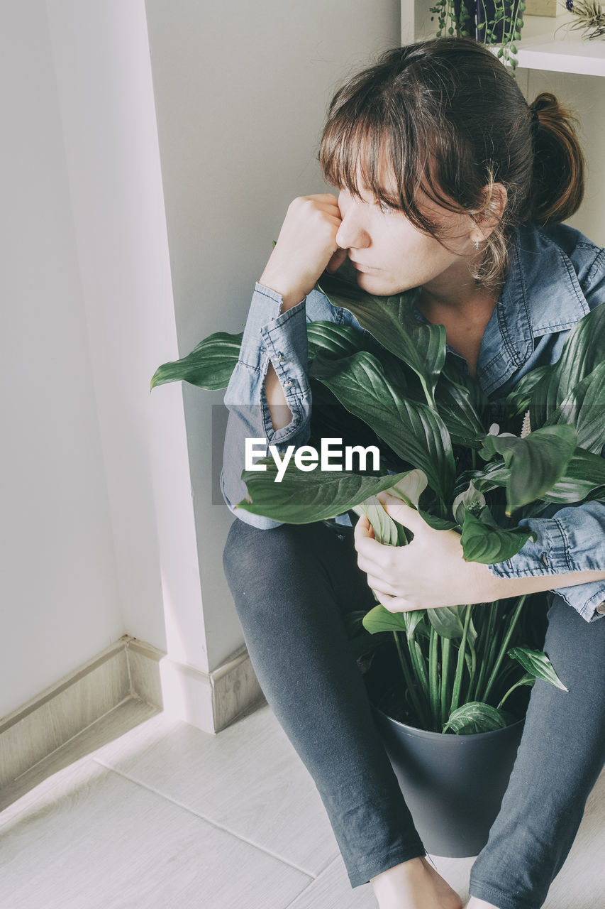Portrait of a woman with brown hair sits on the floor of her house and hugs a plant from a pot