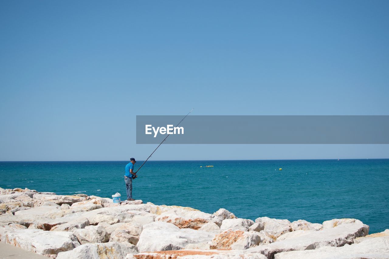 Rear view of man fishing by sea against clear blue sky