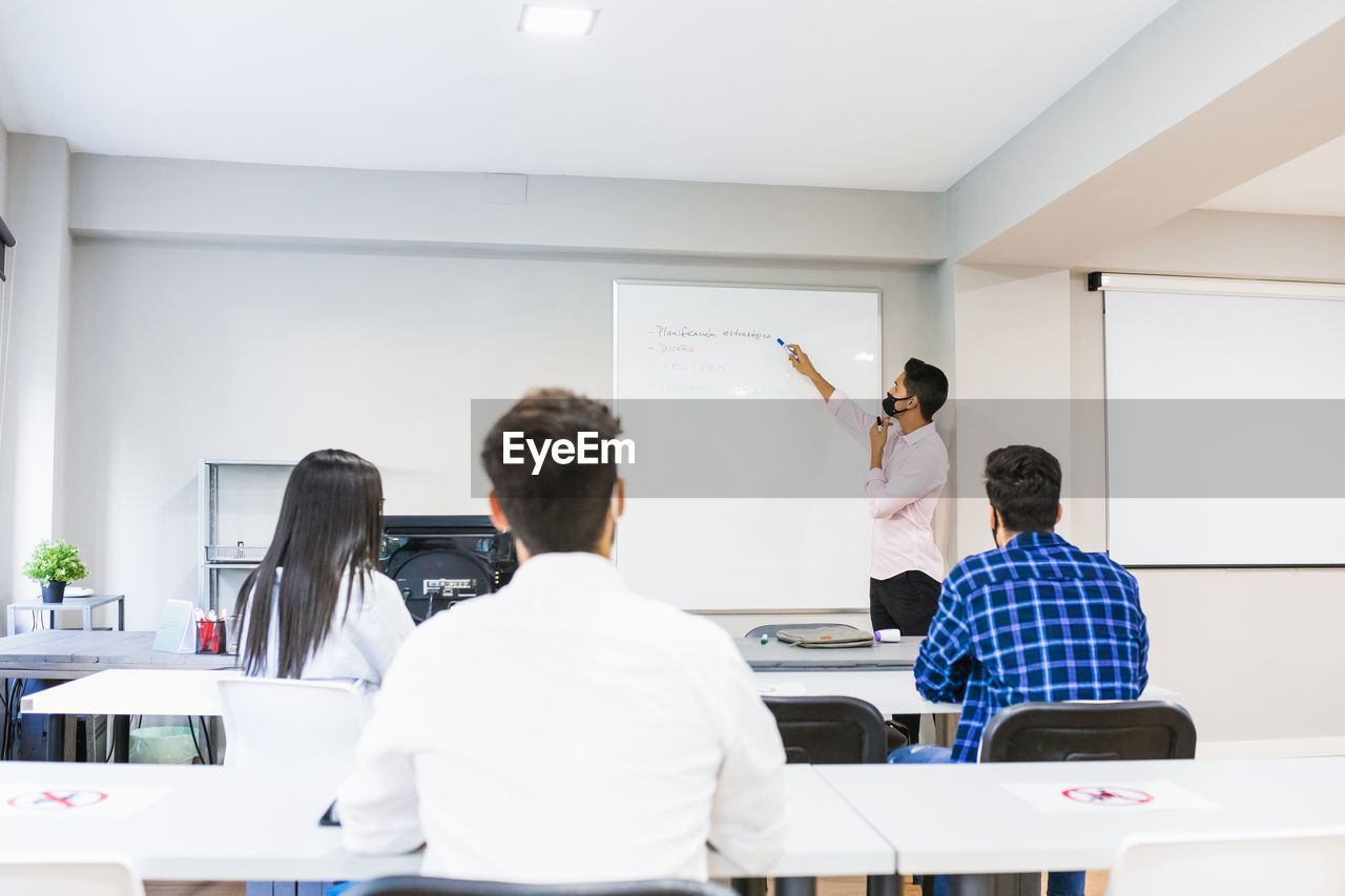 Unrecognizable entrepreneur in mask indicating at board with marker while explaining project to coworkers sitting at tables in office