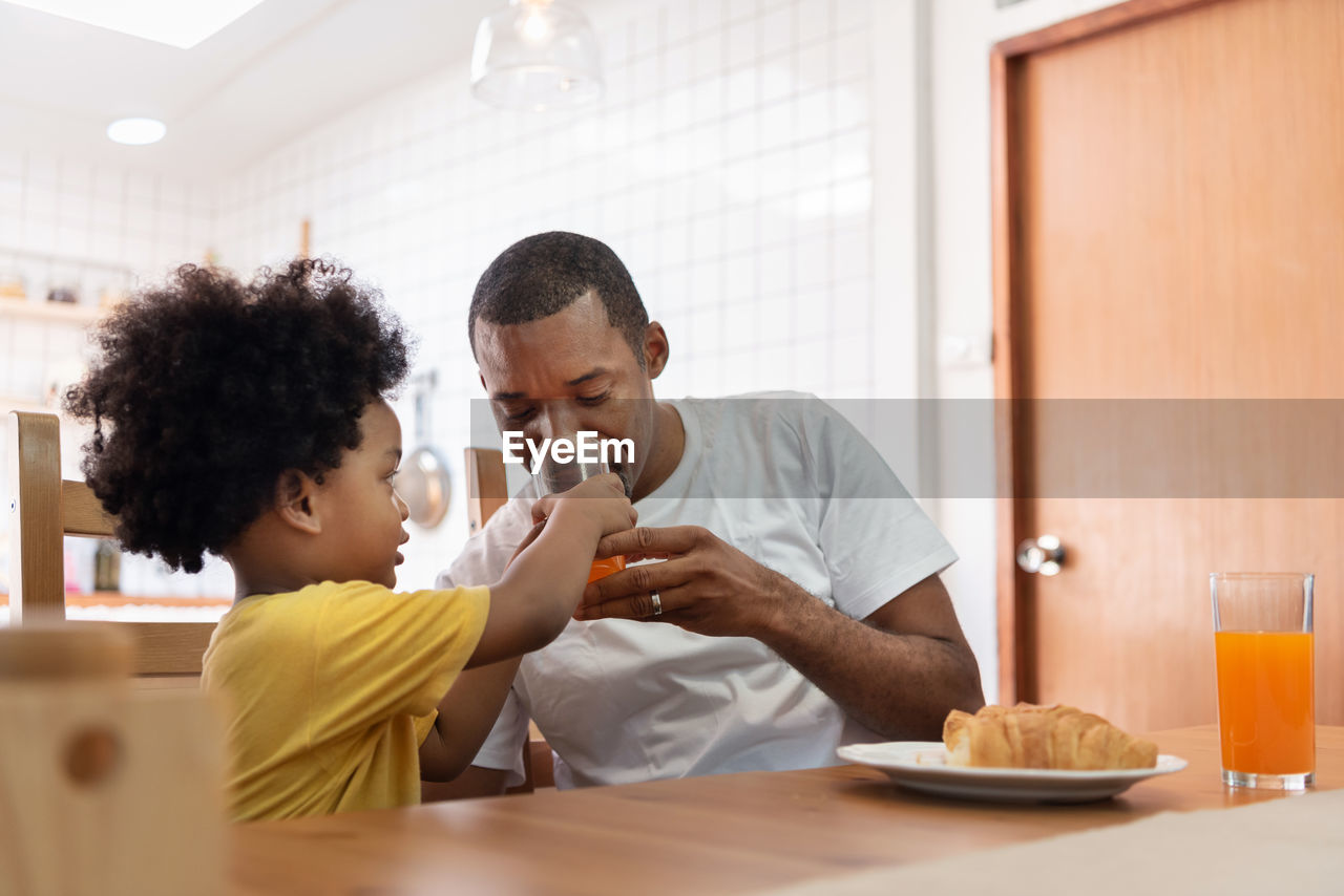 Boy feeding drink to father while having breakfast