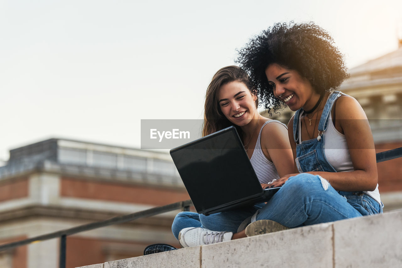 Female friends using laptop while sitting on retaining wall