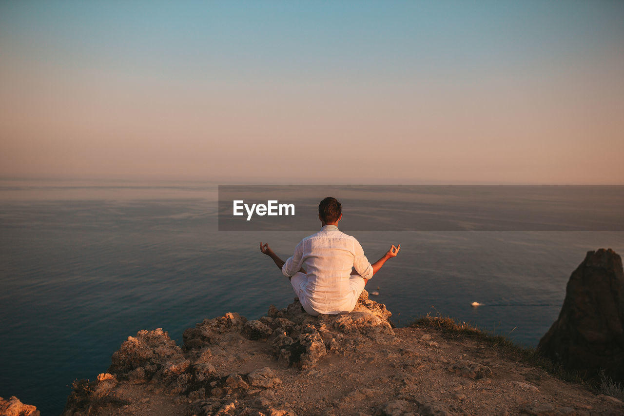 REAR VIEW OF MAN SITTING ON ROCK AGAINST SEA