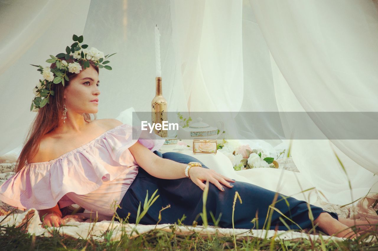 Thoughtful woman wearing wreath resting on field by tent in park