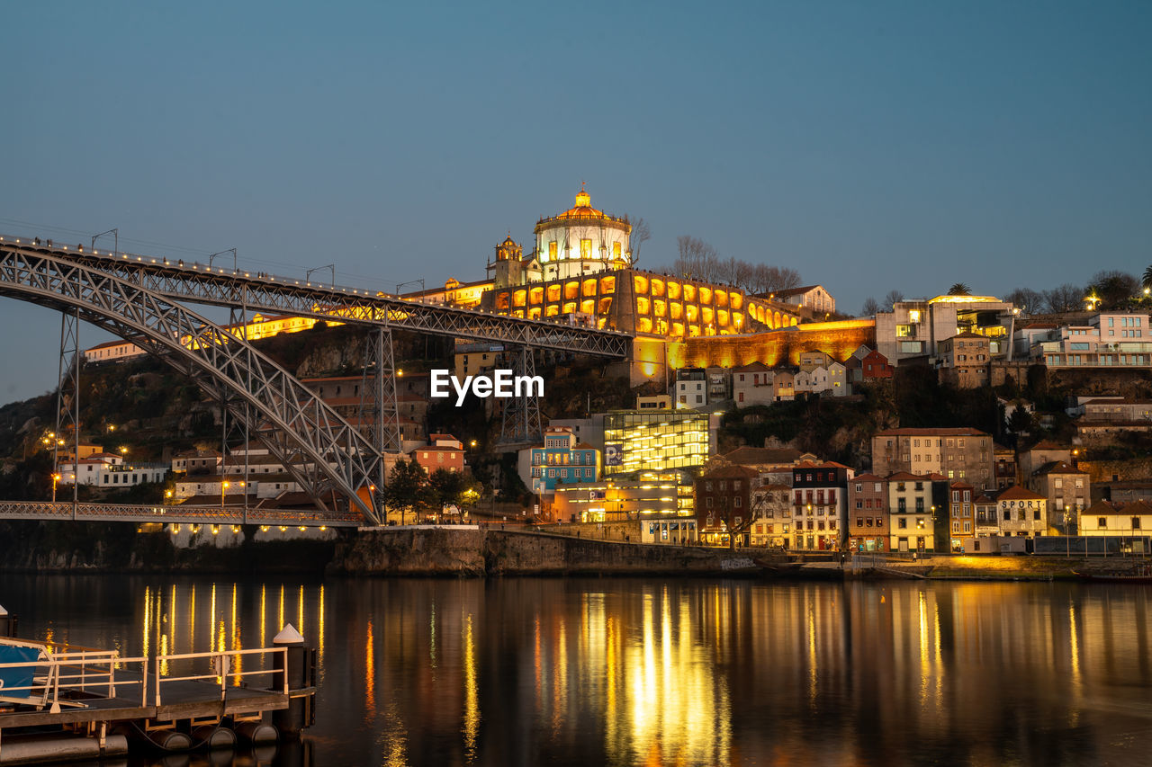 ILLUMINATED BRIDGE OVER RIVER BY BUILDINGS AGAINST SKY IN CITY