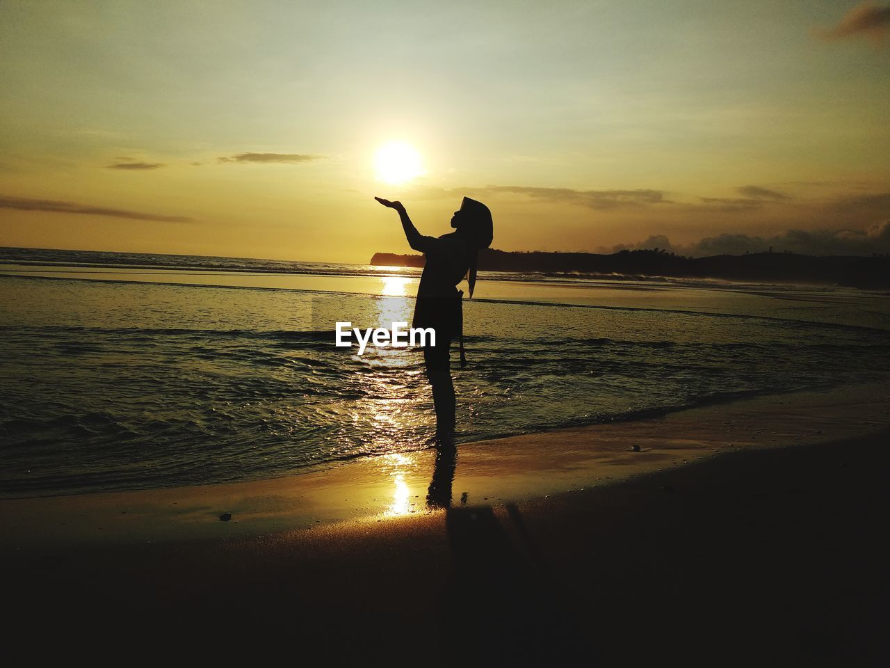 Optical illusion of silhouette woman holding sun while standing at beach during sunset