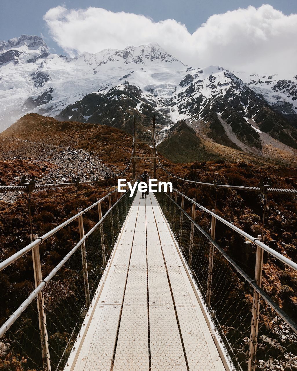 Young man standing on footbridge against snowcapped mountain
