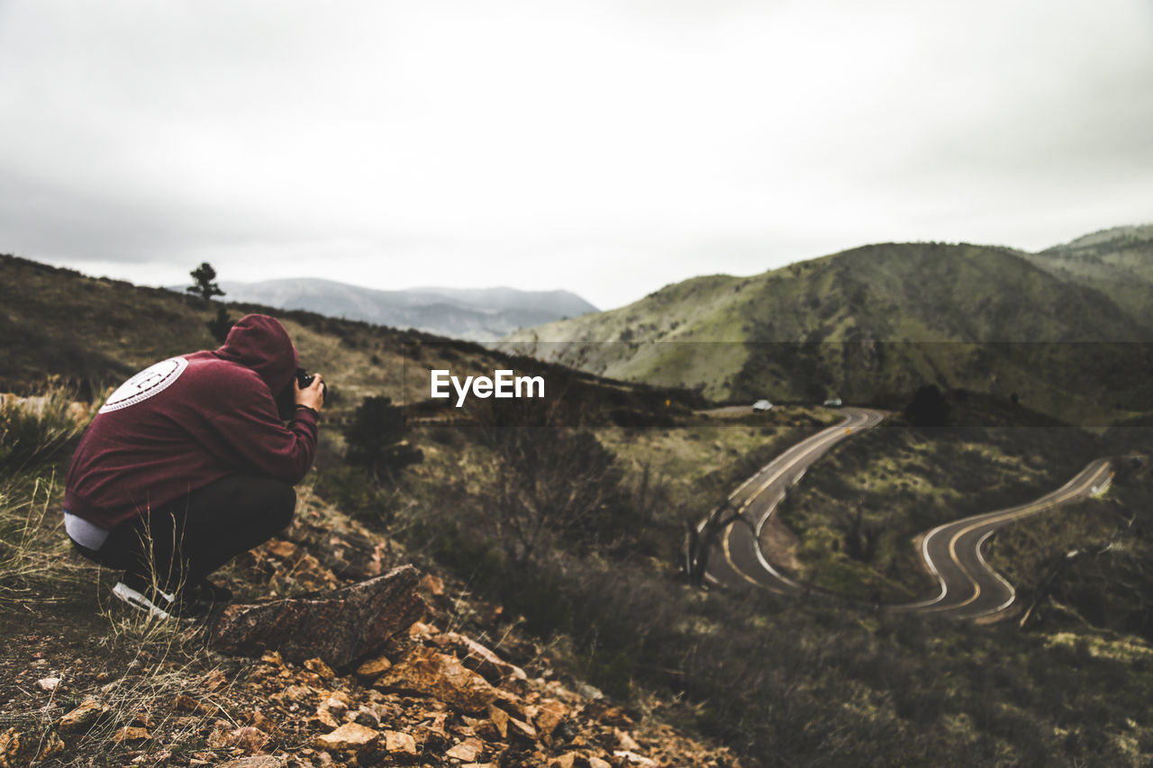 Side view of hiker photographing road while crouching on mountain against sky