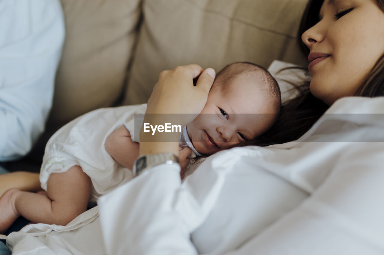 Happy newborn baby boy looking at the camera on top of his mother lying on the sofa