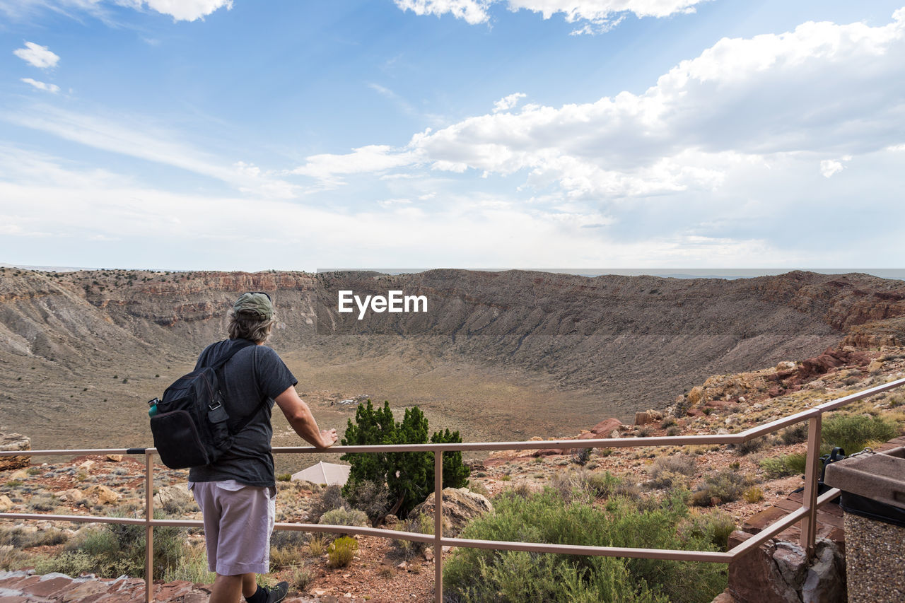 REAR VIEW OF MAN STANDING ON DESERT AGAINST MOUNTAINS