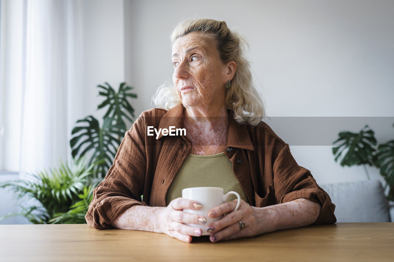 Worried senior woman holding mug sitting at home