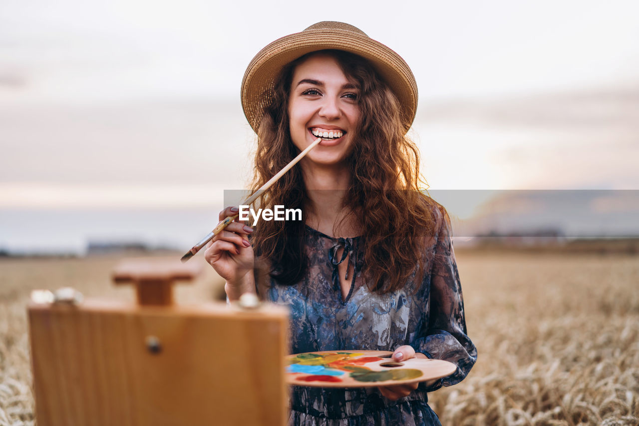 A young woman with curly hair and wearing a hat is painting in nature. 