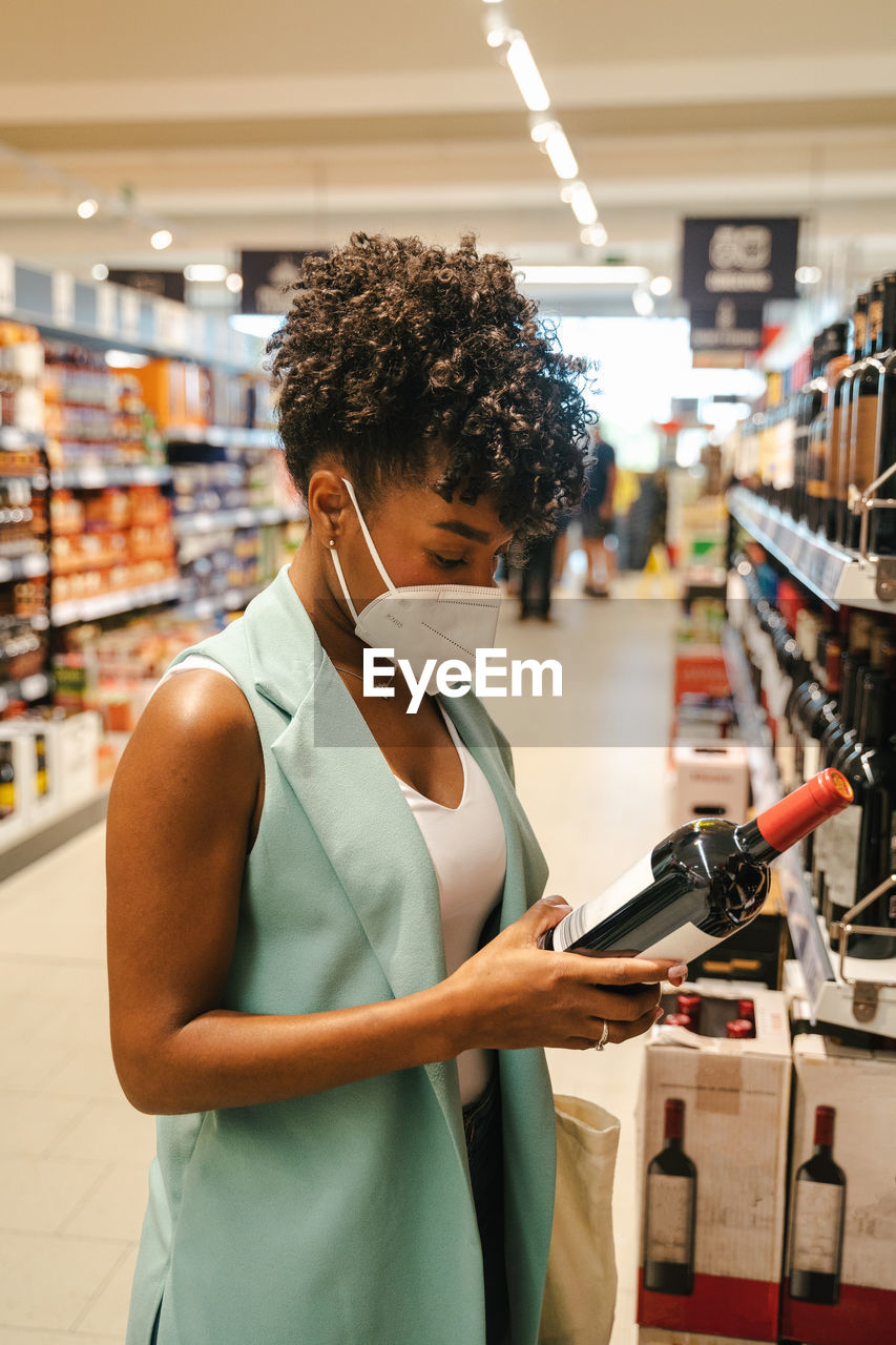 Side view of crop african american female customer in protective mask for coronavirus prevention examining bottle of wine while choosing drinks in supermarket