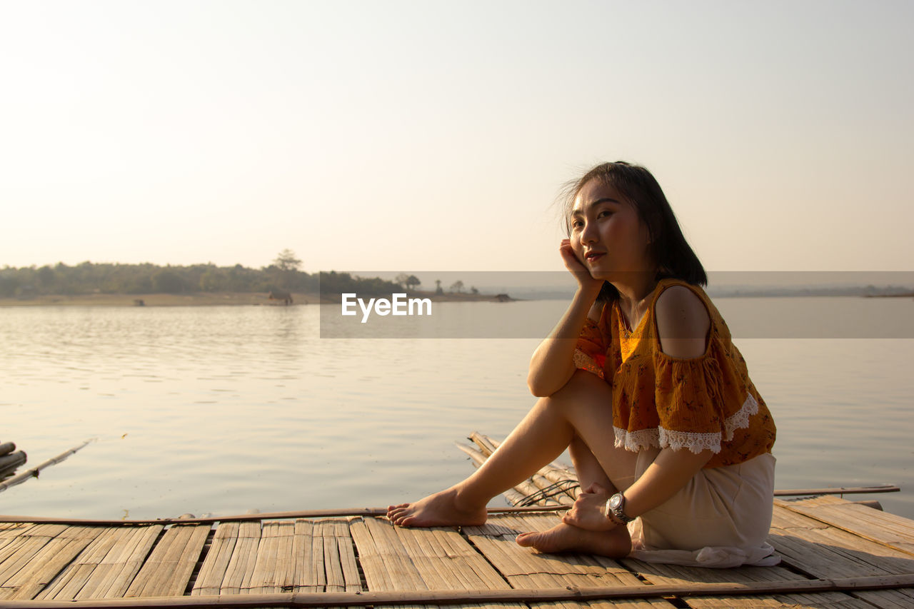 Portrait of woman sitting on wooden raft in lake against clear sky during sunset