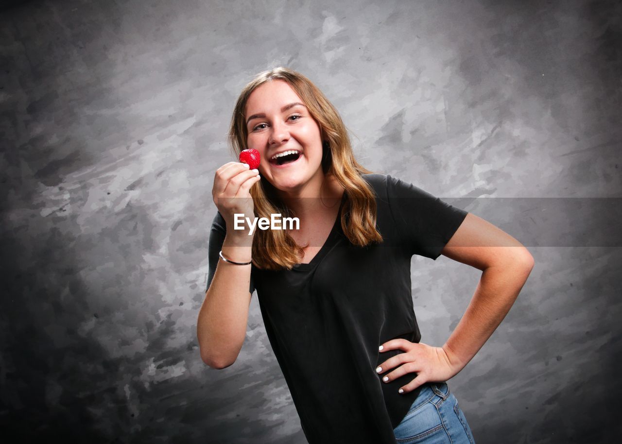 Portrait of laughing woman holding strawberry while standing against wall