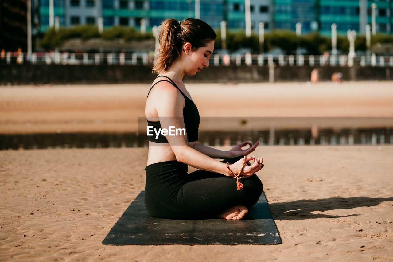 Side view of tranquil female in sportswear sitting on mat in padmasana with mudra hands and meditating while practicing yoga with closed eyes on beach