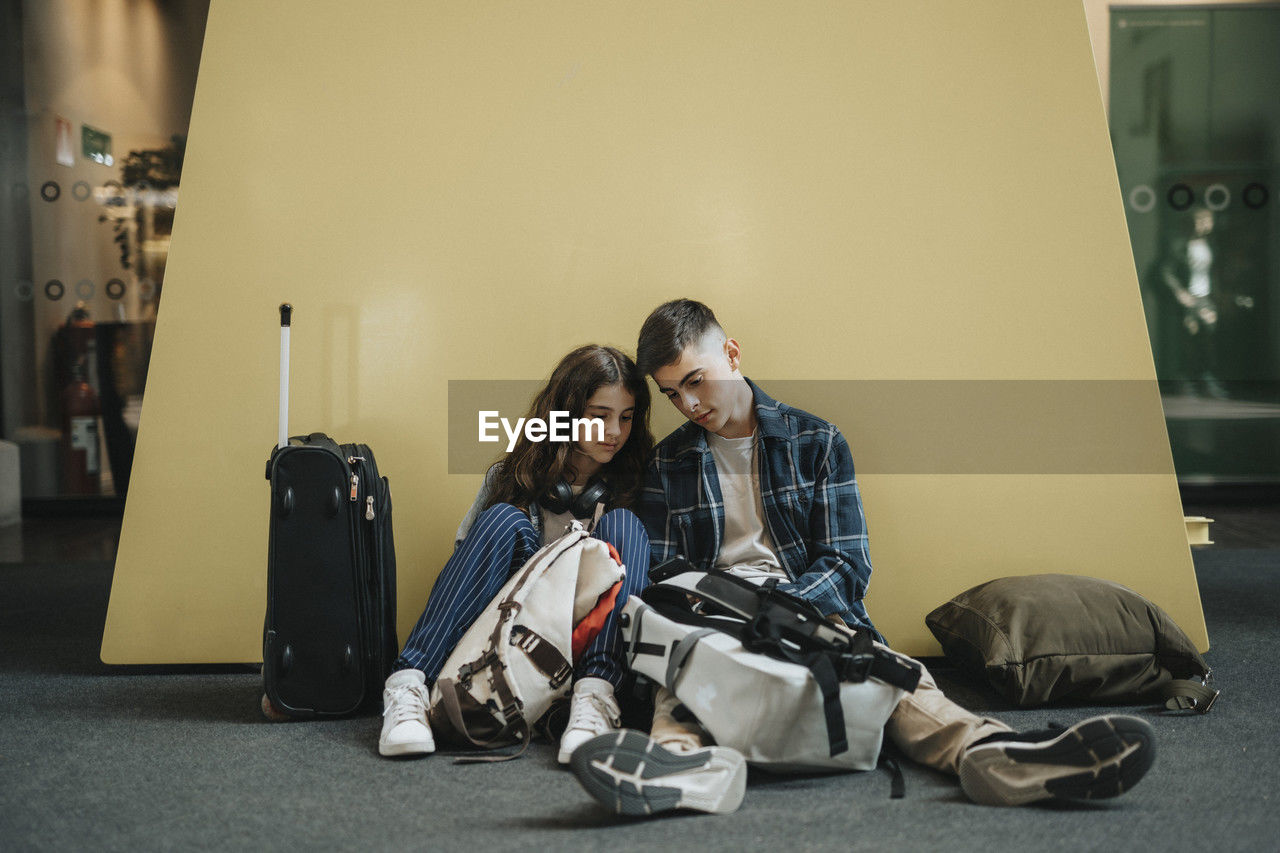 Boy and girl sharing smart phone while sitting with luggage against yellow wall