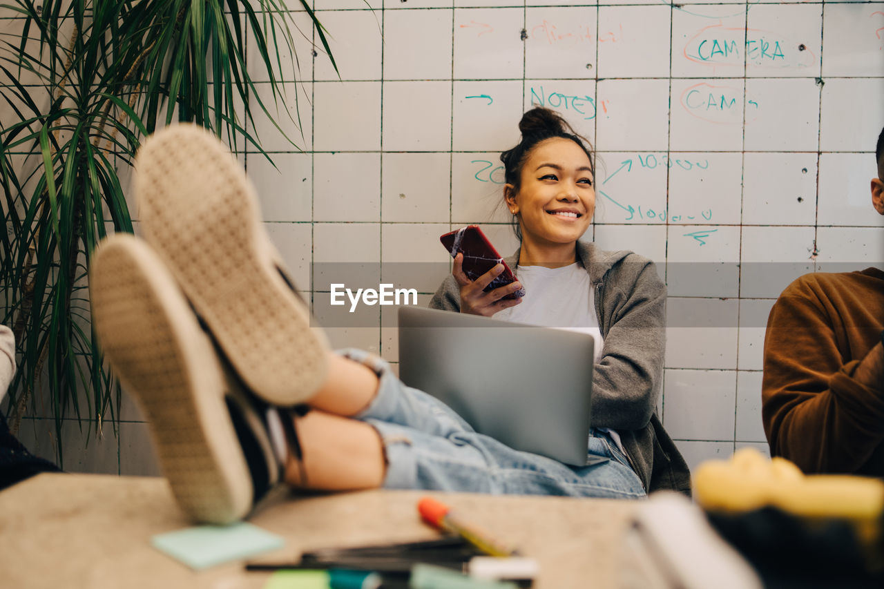 Smiling young businesswoman sitting with feet up on desk using wireless technologies by colleague against wall at office