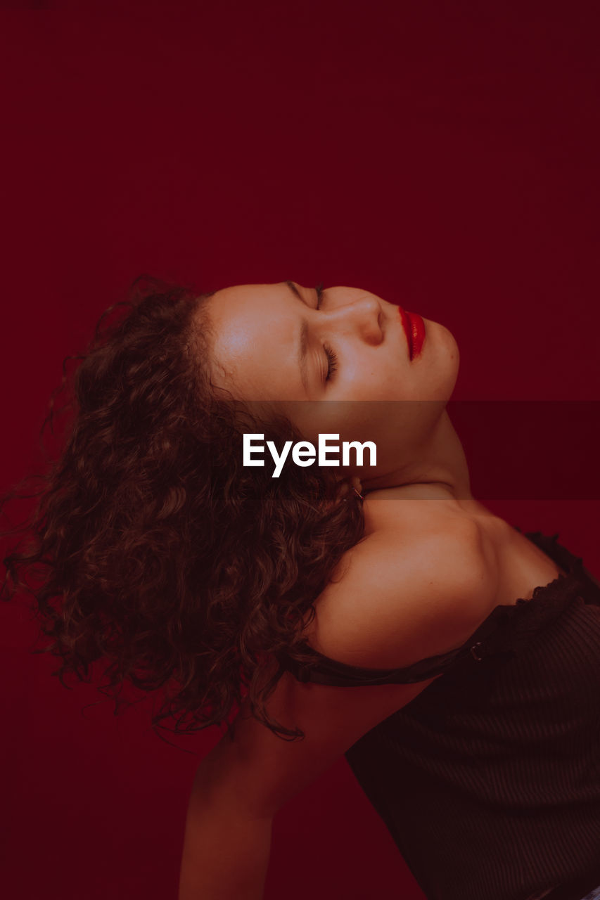 Side view of young woman with eyes closed sitting against red background