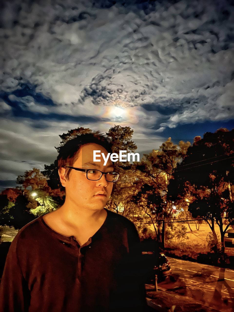 Portrait of young man against cloudy sky, rainbow colored lunar halo and trees at night.