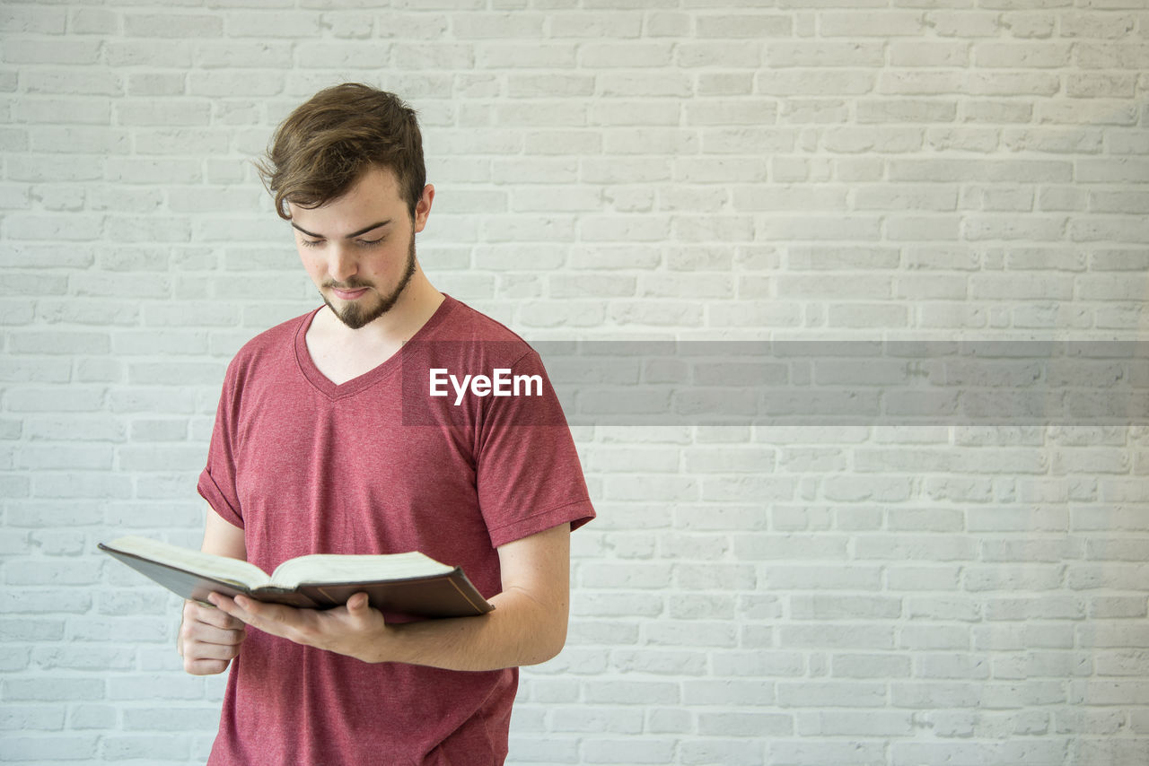 Young man reading bible while standing against brick wall