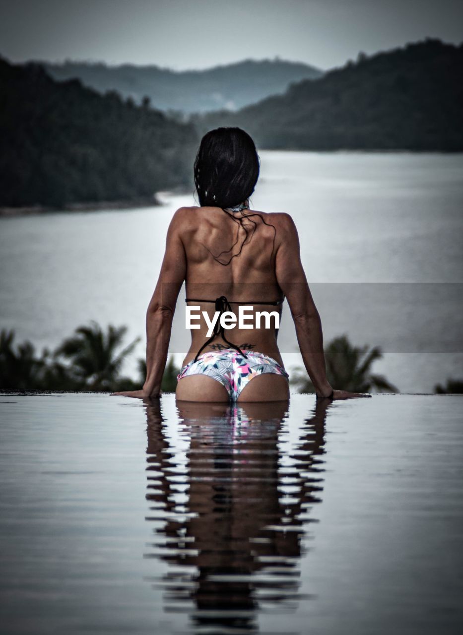Rear view of muscular woman wearing bikini in infinity pool against lake