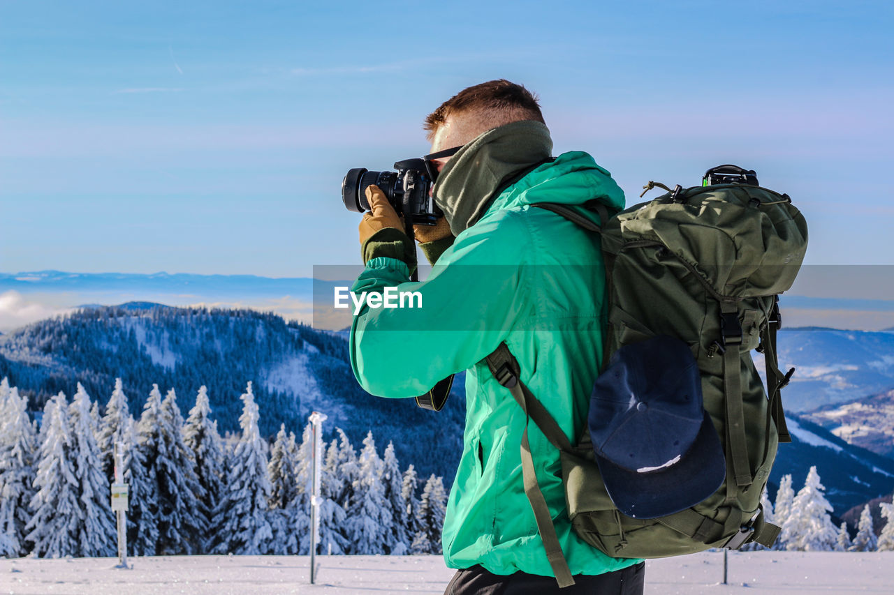 REAR VIEW OF MAN PHOTOGRAPHING ON SNOW COVERED MOUNTAINS