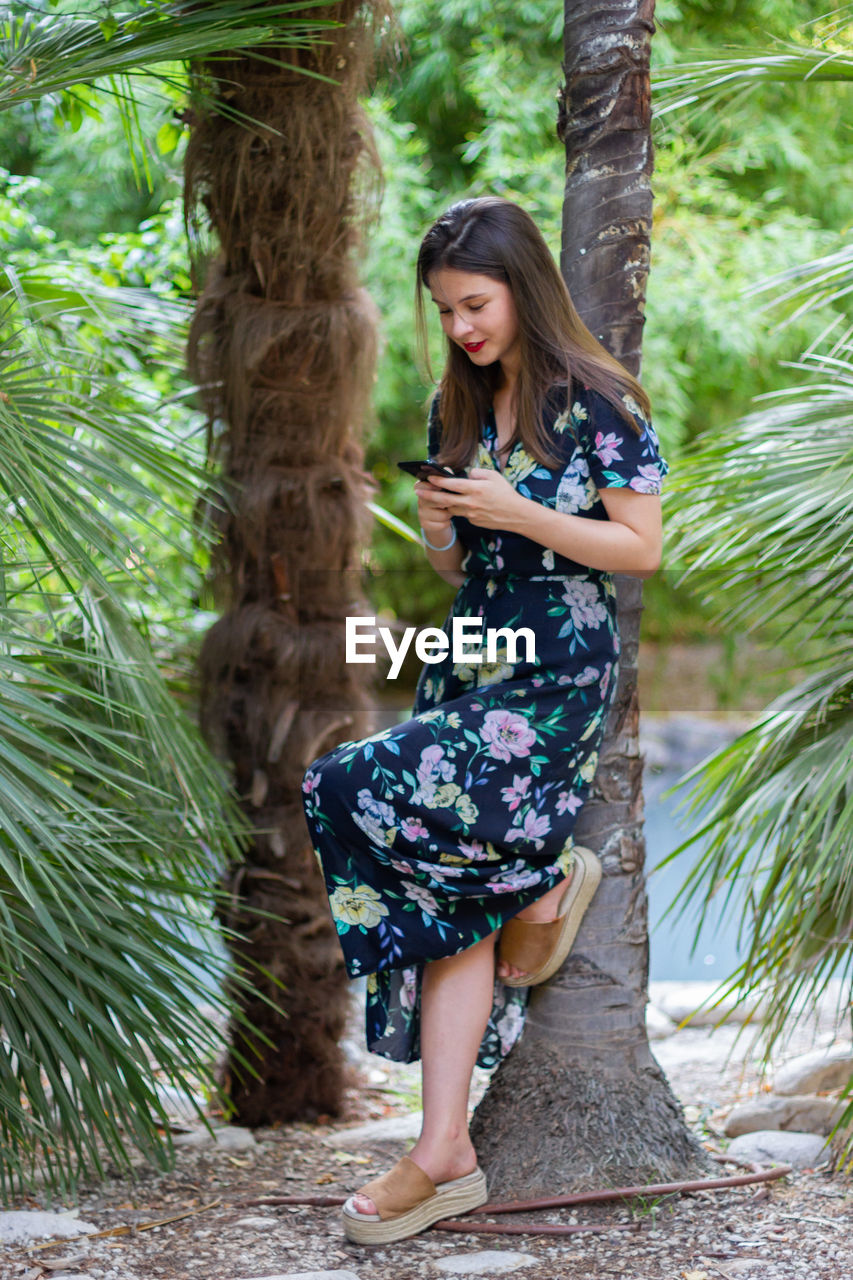 Full length of young woman using mobile phone while standing against trees