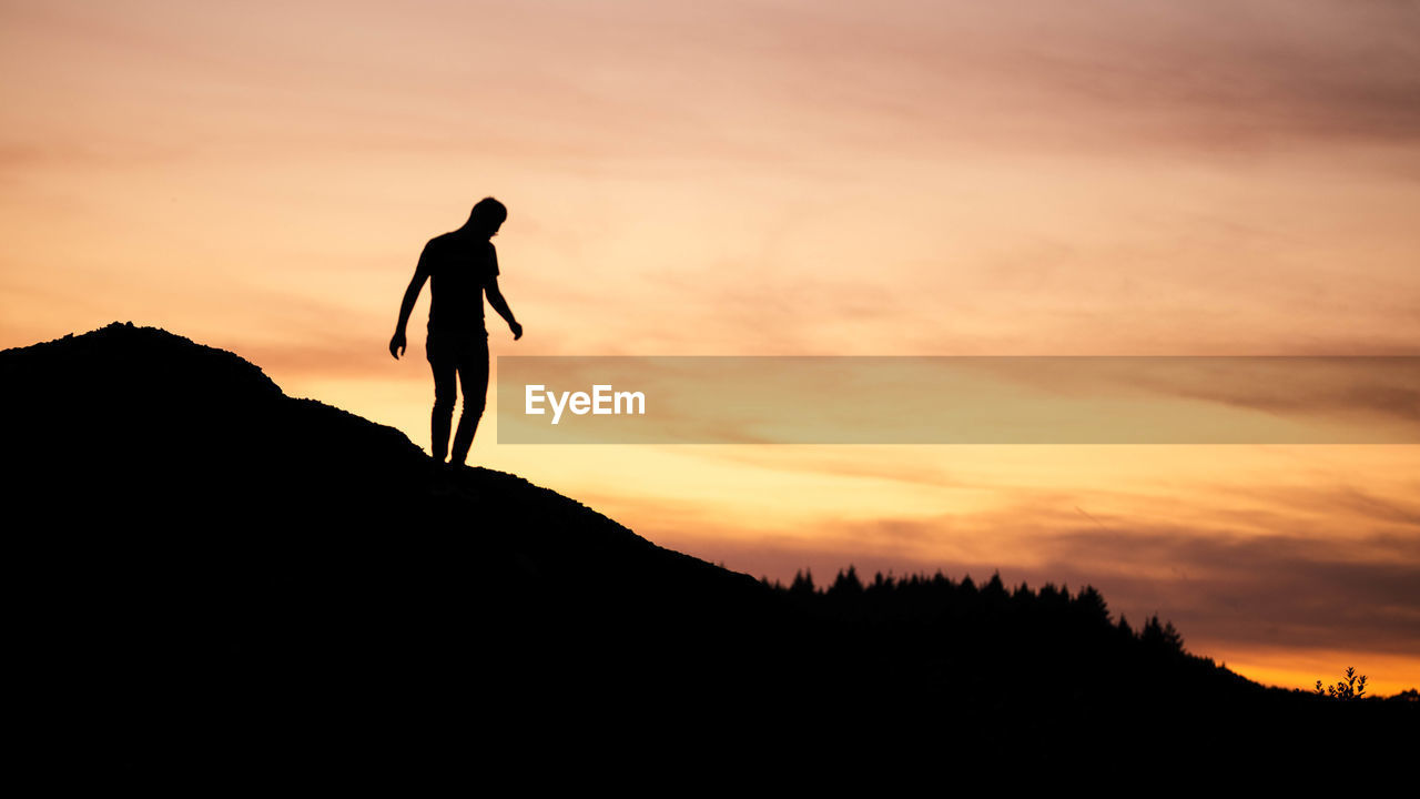 SILHOUETTE MAN STANDING ON MOUNTAIN AGAINST ORANGE SUNSET SKY