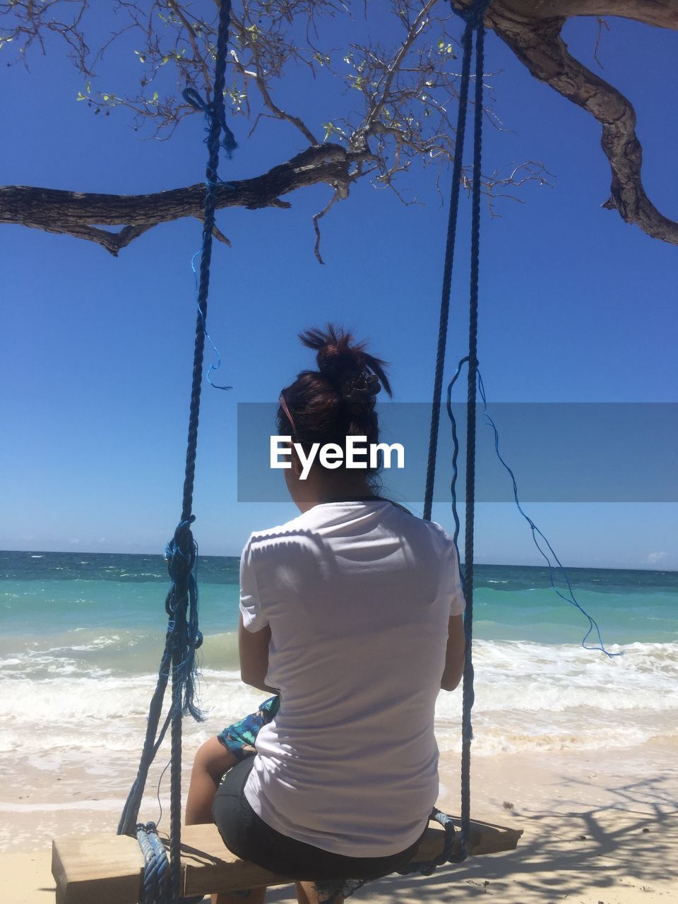 REAR VIEW OF WOMAN SITTING AT BEACH AGAINST SKY