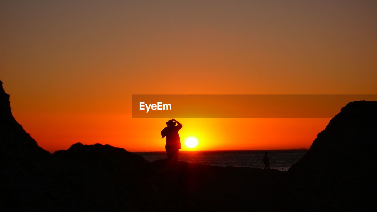 SILHOUETTE MAN STANDING ON SEA AGAINST ORANGE SKY