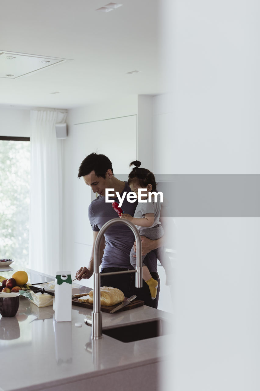 Father carrying male toddler while preparing food in kitchen